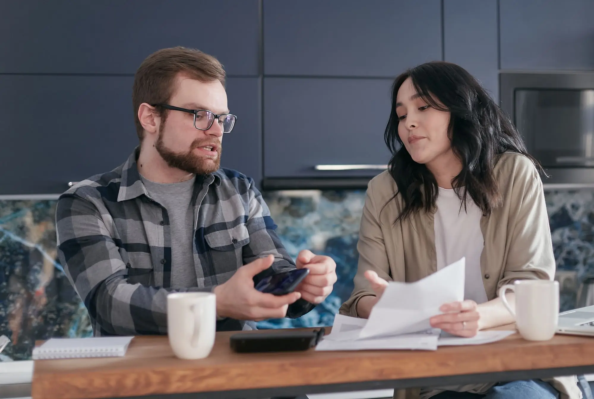 Two business partners at their kitchen bench going over an ATO letter and checking the account balance on a phone